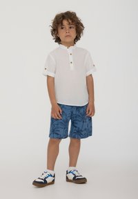 Camicia bianca in lino con colletto alla coreana e polsini abbottonati, abbinata a pantaloni corti blu con motivo di palme. Sneakers bianche e navy.