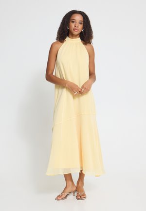 Vrouw met krullend haar die een mouwloze lichtgele halterjurk en open-toe hakken sandalen draagt, staand tegen een witte achtergrond.