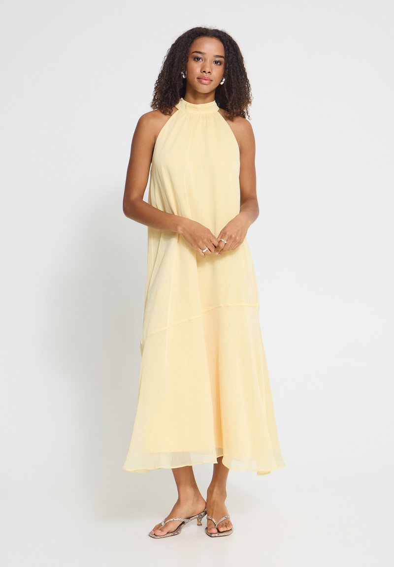Femme aux cheveux bouclés portant une robe sans manches jaune pâle à col halter et des sandales à talons ouvertes, debout sur un fond blanc.