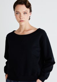 Uma jovem com cabelo curto em ondas usa um sweatshirt preto largo e jeans azuis, em frente a um fundo branco liso.