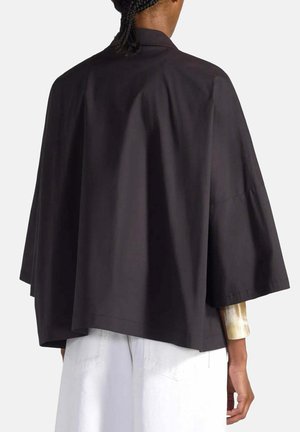 Personne aux cheveux tressés portant un haut noir ample à manches courtes et un pantalon blanc, vue de dos sur un fond uni.
