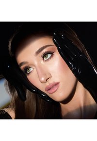 Close-up of a face framed by black glossy gloves. features green eyes, defined brows, full lashes, and soft pink lips against a dark background.