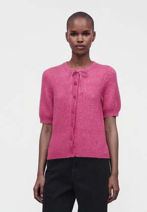 Woman wearing a pink fuzzy short-sleeve button-up cardigan with a tied bow at the neckline and black pants on a plain background.