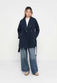 Manteau bleu marine avec un large col, des poches latérales et une ceinture à nouer. Le modèle porte un jean léger et ample bleu clair et des chaussures à motifs.