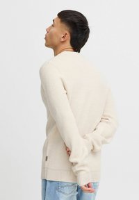 Beige gebreide trui met een geribbelde textuur, ronde hals en lange mouwen. Heeft een zijsplit en een subtiele logobezorging aan de zoom.