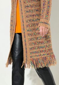 Gilet en tricot beige avec des éclats multicolores, présentant des bords à franges et un design ouvert sur le devant. Associé à un pantalon en cuir noir.