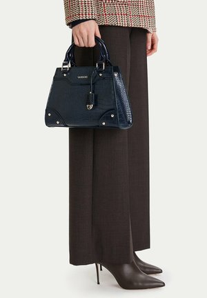Donna con pantaloni a gamba larga marrone scuro e giacca a quadri che tiene una borsa blu navy con texture coccodrillo e indossa scarpe con tacco a punta marrone scuro.