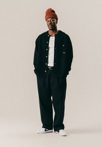 Man in een roestkleurige beanie, zwarte jas, wit shirt, zwarte broek en witte Nike sneakers, staande tegen een effen beige achtergrond.