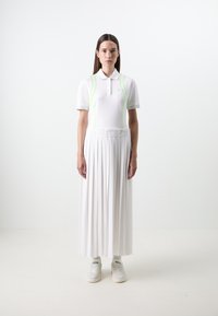 Polo bianco con colletto e dettagli verde neon, abbinato a una gonna bianca a pieghe lunga fino al pavimento. Indossato con scarpe da ginnastica bianche e calze.