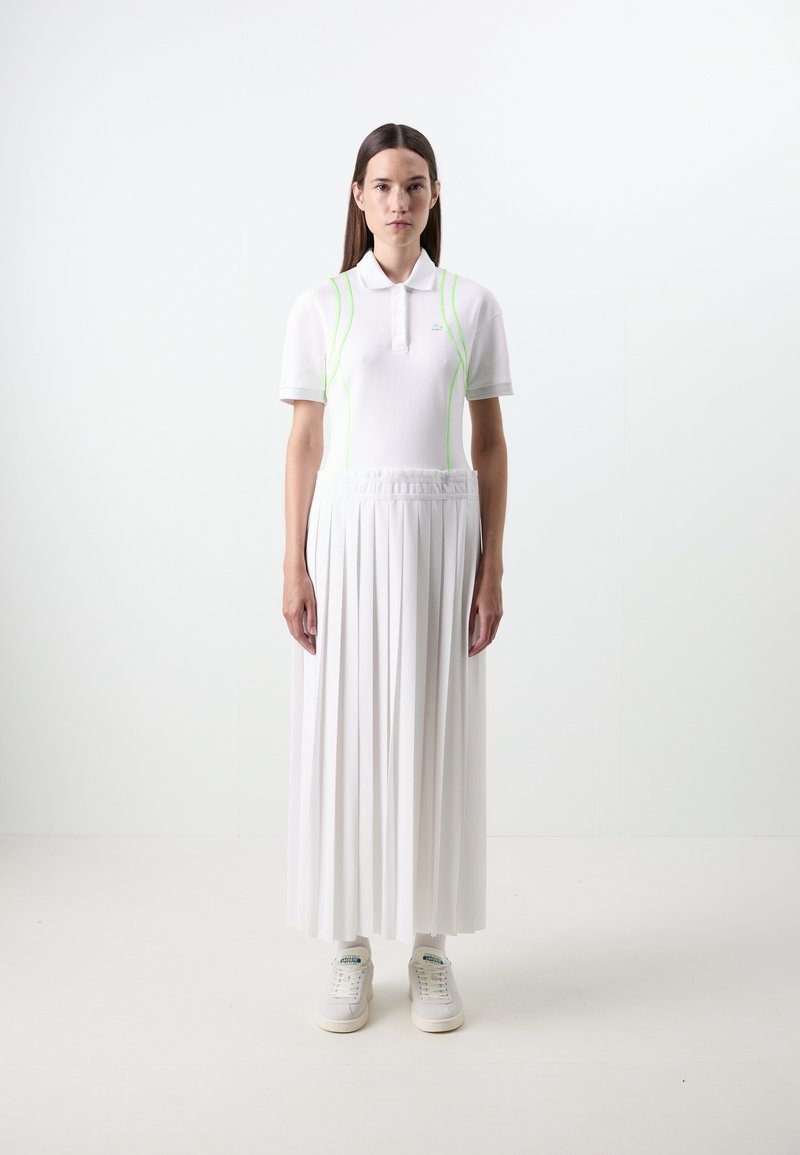 Polo bianco con colletto e dettagli verde neon, abbinato a una gonna bianca a pieghe lunga fino al pavimento. Indossato con scarpe da ginnastica bianche e calze.