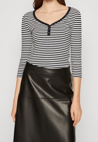 Pitkähihainen, V-pääntiellä varustettu paita musta-valkoisilla vaakasuorilla raidoilla, jossa on kolme nappia kauluksessa. Yhdistetty mustan nahkahameen kanssa.