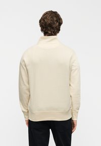 Φυστικί φούτερ με υψηλό γιακά, με λεία υφή και ριμπ μανσέτες. Απλή πλάτη χωρίς ορατά μοτίβα ή διακοσμητικά στοιχεία.