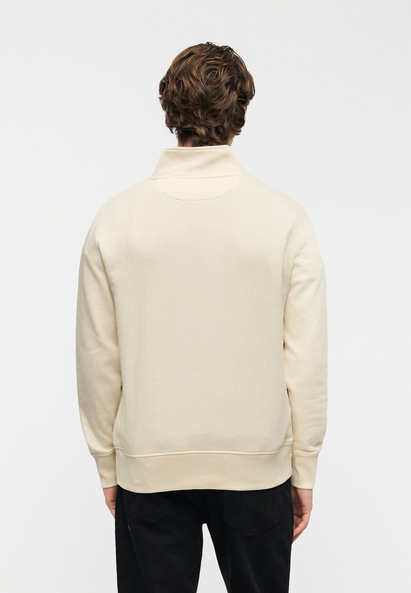 Felpa color crema con collo alto, caratterizzata da una texture liscia e polsini a costine. Schiena semplice senza motivi o dettagli visibili.