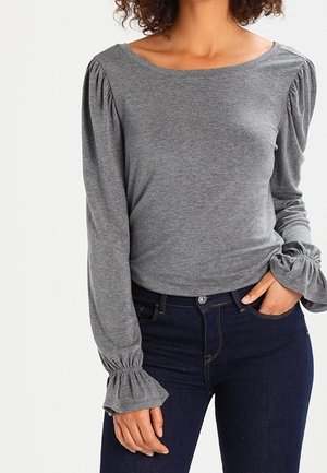 Frau trägt ein graues Langarmshirt mit gepufften Schultern und Rüschen an den Manschetten, kombiniert mit einer dunkelblauen Jeans, die Hand in der Tasche.