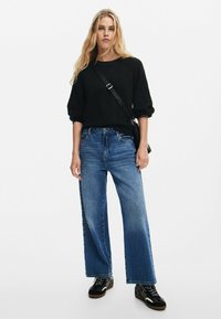 Maglione lungo a maniche lunghe di colore nero e texture, abbinato a jeans blu a vita alta e gamba larga. Sneakers nere con dettagli bianchi e una borsa a tracolla.