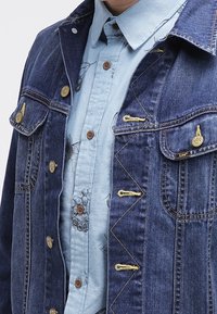 Man in een lichtblauwe bloemenoverhemd met knopen onder een donkerblauwe Lee spijkerjack met messing knopen en gele stiksels.