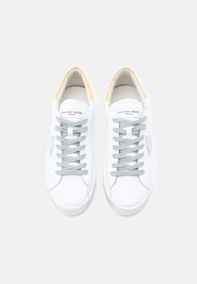 Λευκά δερμάτινα αθλητικά παπούτσια με γκρι κορδόνια και κίτρινη λεπτομέρεια στη φτέρνα. Ομαλή υφή με ελάχιστο λογότυπο στη γλώσσα.