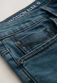 Jean en denim avec un lavage bleu moyen, présentant des coutures contrastantes, une poche avant avec un bouton noir et une étiquette "MOTION FLEX" sur la ceinture.