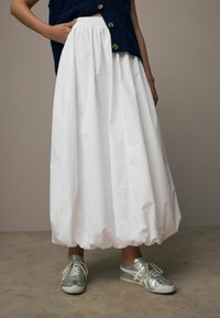 Gonna bianca a palloncino con vita arricciata, abbinata a sneakers argento. Il tessuto è liscio e leggero, creando una silhouette fluida.