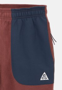 Hnedé a námornícke športové nohavice s jemnou textúrou, vybavené elastickým pásom a trojuholníkovým logom na pravom dolnom vrecku.