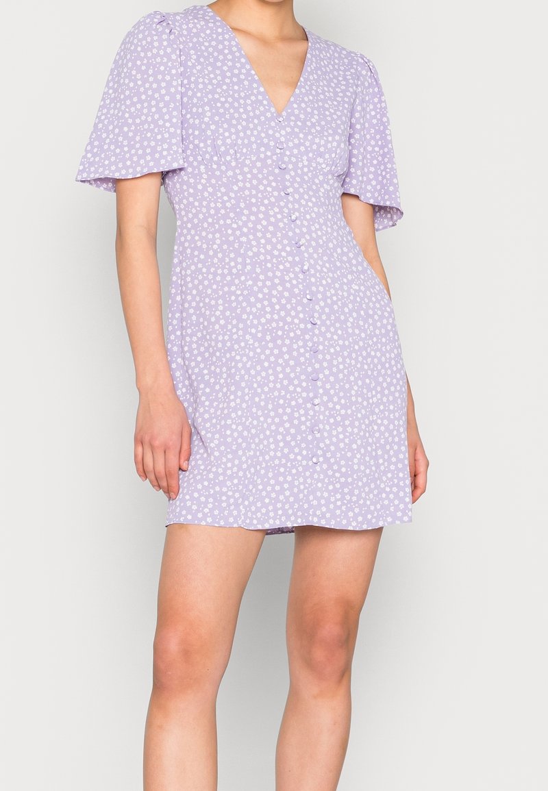 Femme portant une robe courte lavande avec un motif floral blanc, des manches bouffantes, un col en V et des boutons sur le devant.
