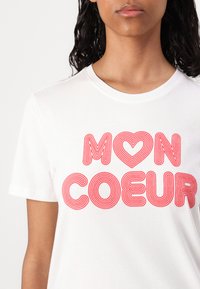 Balta medvilninė marškinė su raudonu kreiviu tekstu "MON COEUR" ir širdelės forma, apvaliu kaklu ir trumpomis rankovėmis.