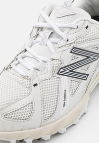 Gros plan sur une chaussure de sport blanche New Balance avec tissu en mesh, superpositions synthétiques, lacets blancs et grand logo "N" à contour noir sur le côté.