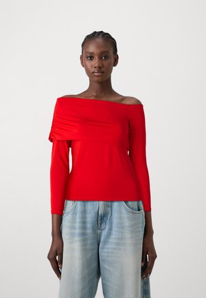 Top rosso senza spalle con maniche lunghe aderenti e dettagli arricciati sul petto. Abbinato a jeans azzurri a vita alta e larghi.