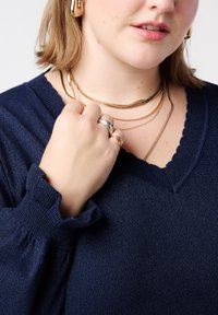 Marineblauwe trui met een geschulpte halslijn, gecombineerd met meerdere goud- en zilverkleurige gelaagde kettingen. Heeft een detail van gerimpelde mouwen.