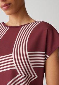 Burgunderfarbenes Oberteil mit kurzen Ärmeln, das weiße diagonale Linien aufweist, die ein geometrisches Muster bilden. Glatter Stoff mit einer lockeren Passform.