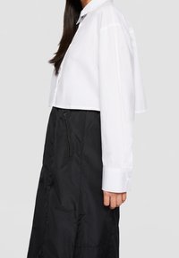 Chemise blanche courte à boutons avec col classique, associée à une jupe noire avec une poche zippée et un tissu texturé.