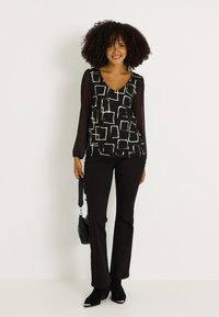 Blusa negra con un estampado geométrico blanco, con mangas largas transparentes. Combinada con pantalones acampanados negros y botas de tobillo negras.