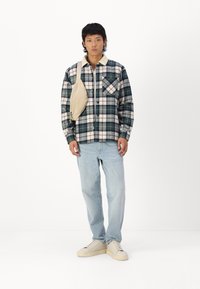 Chemise à carreaux verte et crème, avec un col et des poches poitrine, associée à un jean bleu clair et des baskets ivoire, portant un sac à bandoulière beige.