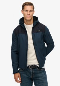 Marineblaue Softshelljacke mit Kapuze, matter Oberfläche, kontrastierende schwarze Schultern und seitlichen Reißverschlusstaschen; getragen über einem hellen Hemd.