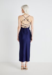Navy satijnen jurk met een aansluitende silhouet, voorzien van een crisscross rugontwerp en strikdetails, aangevuld met sleehakken.