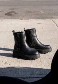 Paar schwarze Leder-Stiefeletten mit klobigen Sohlen und seitlichen Reißverschlüssen, die auf einem Betongehweg im Sonnenlicht stehen.