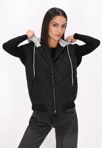 Gilet trapuntato nero con cappuccio grigio, zip frontale e orlo a costine. Il modello sistema il cappuccio mentre indossa una maglietta nera a maniche lunghe e jeans.