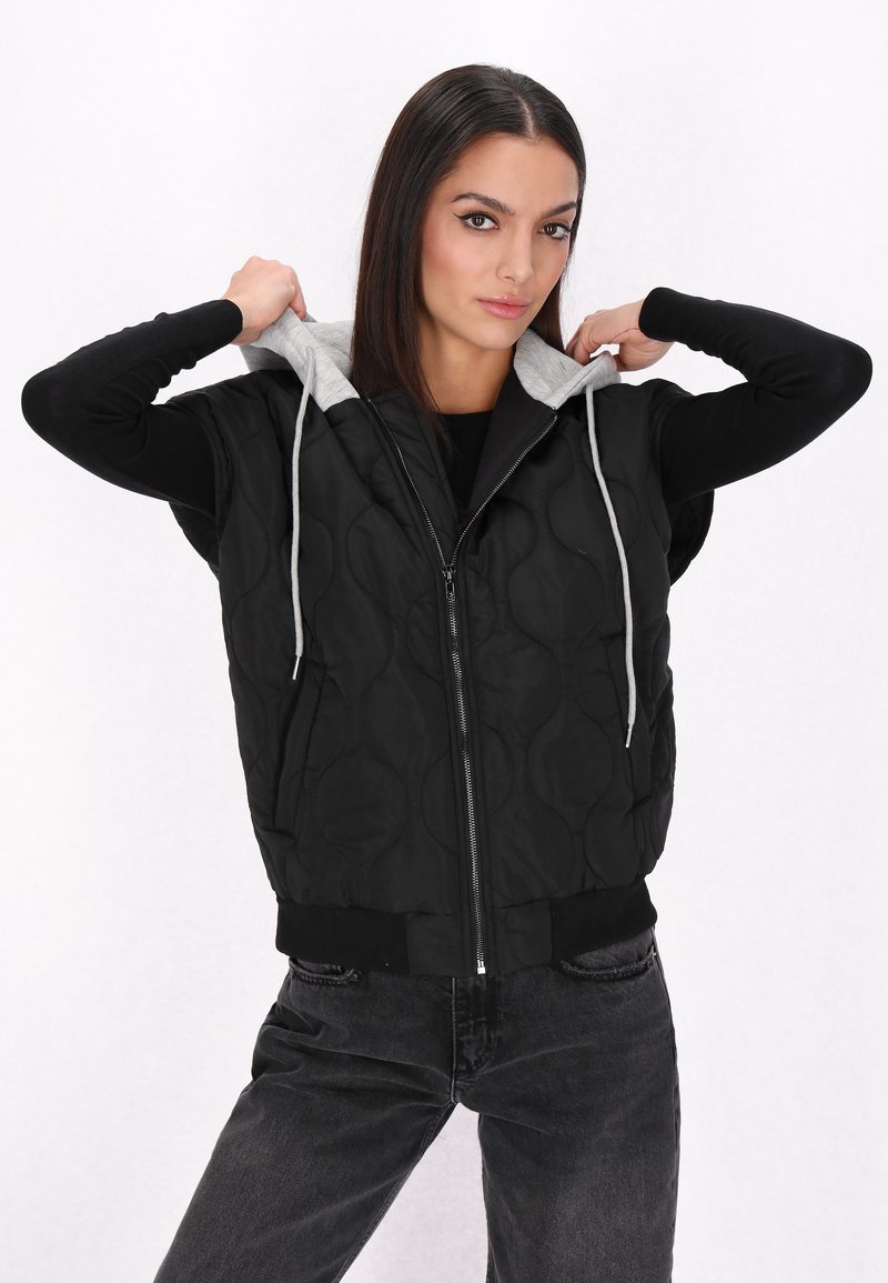 Gilet trapuntato nero con cappuccio grigio, zip frontale e orlo a costine. Il modello sistema il cappuccio mentre indossa una maglietta nera a maniche lunghe e jeans.