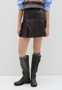Bruine faux leren geplooide rok met gespsdetails, gecombineerd met bruin kniehoge laarzen en grijze kniekousen.