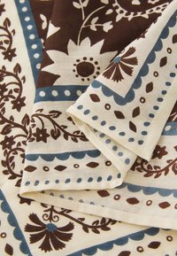 Baumwollstoff mit einem floralen und geometrischen Muster, das braune und blaue Akzente auf einem cremefarbenen Hintergrund zeigt. Gefaltete Kanten sind sichtbar.