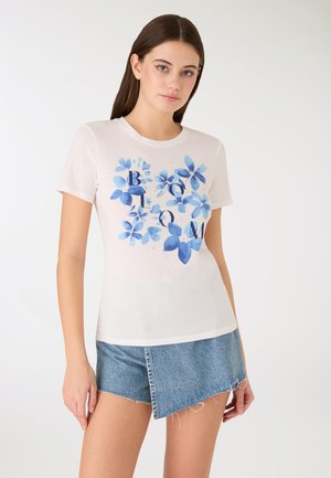 Giovane donna che indossa una t-shirt bianca con stampa floreale blu e la parola "BLOOM", abbinata a pantaloncini di jeans asimmetrici.