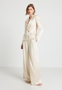 Cremefarbener Satin-Doppelreiher-Blazer mit goldenen Knöpfen, kombiniert mit Satin-Hosen in weitem Schnitt. Glatte Textur und maßgeschneiderte Passform.