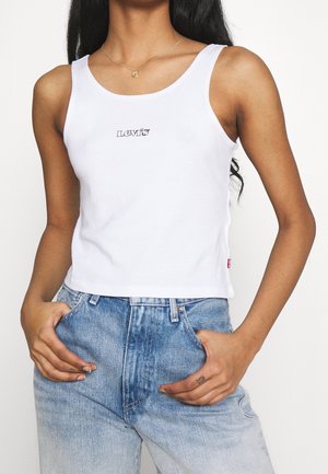 Femme portant un débardeur blanc sans manches Levi's et un jean taille haute bleu clair, les mains dans les poches, sur un fond uni.