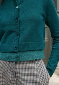 Tyrkysový cardigan na knoflíky přes shodný tyrkysový top, doplněný hnědými a černými kostkovanými kalhotami, ruka v kapse.