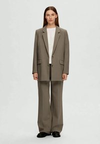 Blazer grigio su misura con due bottoni e tasche a pattina, abbinato a pantaloni larghi coordinati e una t-shirt bianca sotto. Scarpe nere.