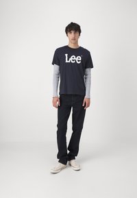 Mladý muž stojaci, oblečený v čiernom tričku Lee cez sivé dlhé rukávy, čierne nohavice a biele tenisky na jednoliatom svetlom pozadí.