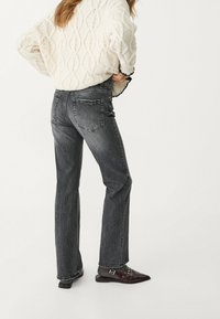 Jeans grigi con taglio dritto, dotati di tasche posteriori e una finitura sbiadita, abbinati a un maglione in maglia beige testurizzata e calzature scure.