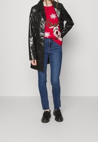 Černý lesklý trenčkot přes červený svetr s motivem soba, doplněný modrými úzkými džínami a černými šněrovacími botami.