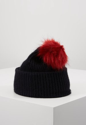 Bonnet noir tricoté avec revers plié, orné d’un grand pompon rouge duveteux, présenté sur une surface blanche.