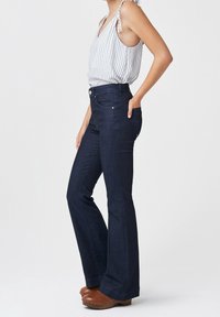 Ausgeflockte dunkelblaue Jeans aus Denim mit hoher Taille, ausgestattet mit Taschen und subtilen Kupferakzenten. Kombiniert mit einem gestreiften Oberteil.